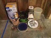Photo of assortment of kitchen appliances and accessories including crockpot, blender, plates, molds, basket with canning supplies and spatulas placed on floor with wood paneled wall background.