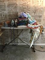 Full view of the ironing board with stacked linens and two irons placed on it.