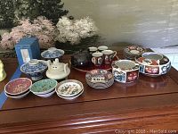 Photo shows assorted Japanese china on a wooden surface including hand-painted teacups and saucers, small plates, rice bowls, ashtrays, teapots, and decorative mugs in a variety of colors and traditional Japanese designs.