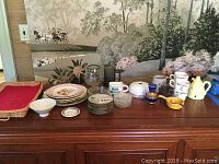 Wide view of entire assortment consisting of ceramic, porcelain dishware, coffee cups, mugs, coasters, glass candle holder, wicker basket, and trays.