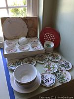 Full view of the dishware lot, showing stacked small bowls (pink and green Kaiser), floral plates laid out, a red decorative plate and a box containing smaller bowls and tea cup.