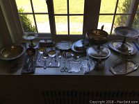 Variety of silver plated serving dishes, utensils, and a sterling silver candle holder arranged on a window ledge.