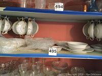 Shelves holding over 20 porcelain tea cups hanging on hooks and stacked porcelain plates in different sizes, visible delicate floral patterns and colored rims