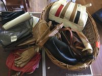 Photo showing a basket containing multiple handbags in various shades of brown, black, red, along with a pair of leather gloves and a woven basket handbag with white and brown stripes.