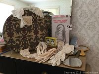 Overview of gloves, lace collars, small round suitcase with floral pattern, boxed brass vanity mirror, and vintage yellow box.