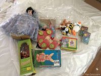 Photo showing multiple children's toys including a doll with purple dress, boxed Hawaii Princess musical doll, assorted beanie babies, light up buttons, and other toys arranged on white surface.