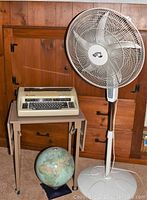 Full view of metal cart with folding sides, Olympia electronic compact typewriter on top, Hampton Bay pedestal fan beside the cart, world globe on the floor.