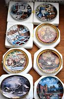 Eight collectible plates from The Bradford Exchange in packaging placed on a wooden surface, showing various designs including cats, interiors, and winter landscapes.