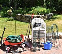 Overview of lot showing red metal wagon carrying garden hoses, stack of white plastic chairs, metal plant supports, watering can, shovel, bird feeders, and mailbox on grass and brick patio.