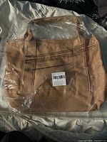 Front view of tan canvas tote bag in plastic wrap showing printed text and handles