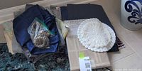 Stack of assorted table linens, placemats, napkin rings in zip bag, crochet doilies, packaged tablecloth visible