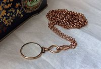 Coiled gold-plated chain with magnifier pendant beside decorative box
