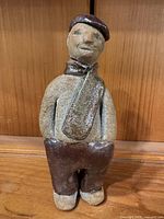 Front view of handmade stoneware folk art figurine showing cap, scarf, trousers, facial features, and overall condition