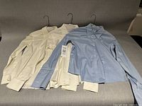 Three long-sleeve tailored shirts on hangers, light blue and cream colors, showing overall condition and style