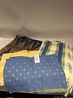 Folded piles of assorted patterned fabrics: blue with gold floral, yellow check, cream plaid, navy with gold motif