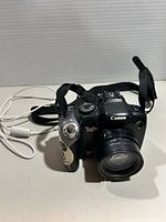 Front angled view of Canon camera with lens cap off, showing attached strap and USB cable