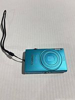 Front view of Canon PowerShot turquoise camera showing lens cover, logo, flash and wrist strap