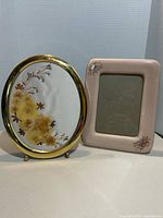 Two picture frames, one oval gold-tone with dried flower art, one pink ceramic rectangular with floral detail