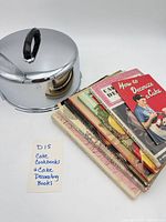 Stack of seven cookbooks beside chrome cake cover