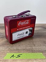 Front of red Coca-Cola tin showing branding, handle and rules card window