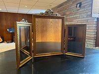 Trifold mirror open on tabletop showing gilded frames and missing right crest ribbon