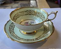 Teacup on saucer, side view showing pale celadon glaze, gilt scroll border and gold foot