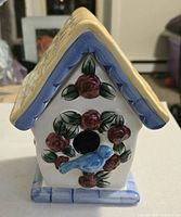 Front view of glazed ceramic birdhouse candle showing roof colors, floral wreath, and blue bird detail