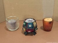 Front view of three candle holders: clear ribbed glass, multicoloured mosaic on stand, amber hobnail