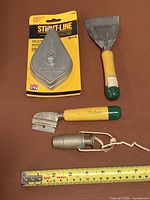 All four items: sealed chalk line reel, large scraper, razor scraper, plumb bob with tape measure for scale