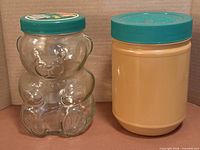 Glass teddy bear jar and plastic jar side by side with lids on