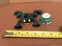 Plush frog and googly-eyes pom figure beside ruler for scale