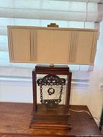 Full view of lamp with shade showing wood base, open frame, metal filigree, chain ornament, pleated shade