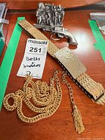 Wide metal belt, chain sash with tassel, pewter figurine and revolver belt buckle arranged on tabletop