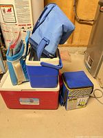 Stacked view of red Coleman cooler, blue hard cooler, two blue insulated bags and Sportcraft darts case
