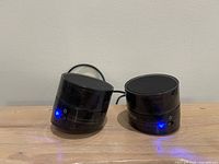 Two black cylindrical speakers on wooden surface, blue LEDs lit
