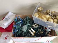 Box containing mixed lights, gold ornaments, bead garland, ribbon, and holiday hand towels