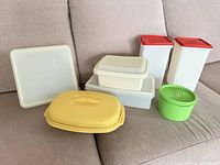 All Tupperware pieces on sofa: two tall containers with red lids, two shallow containers with lids, yellow oval container, green round container, square lid