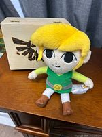 Front view of Link plush seated against wooden box showing Triforce crest