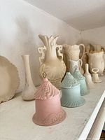 Shelf with multiple cream bisque vases, pitchers, plate, and two pastel bell ornaments
