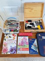 Overall lot view: brushes bin, acrylic paint box, watercolor book, pad, pencils