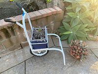 Side view of white metal one-wheeled planter cart, blue wooden planter box with succulents, separate plastic pot of succulents visible