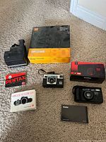 Group shot of all six items: Polaroid Spirit 600, Kodak Carousel slide tray box, Pentax cloth, Minolta manual, Kodak Instamatic X-15, boxed Vivitar ZM85, loose Vivitar camera