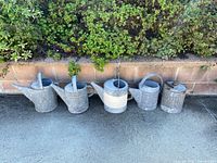 Five galvanized watering cans lined against brick wall