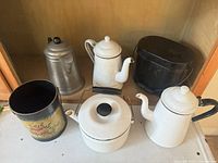 Group shot showing all six items: two white enamel coffee pots, enamel saucepan with lid, aluminum percolator, floral tin, black lidded pail