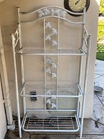 Full view of white metal folding shelf unit showing three shelves and decorative vine motif
