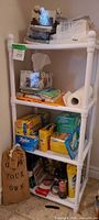Four-tier white plastic shelf loaded with boxed Ziploc, Press’n Seal, Swiffer cloths, paper towel roll, aerosol cleaners, microfibre cloths and other supplies