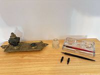 full lot overview on tabletop: brass inkwell tray with lidded inkwell, loose glass bottle, acrylic pen rest, pens and nibs