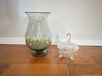 Smoky gray hurricane vase filled with iridescent pebbles beside clear cut glass covered candy dish