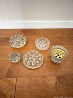 Five glass flower frogs laid out on wood floor, showing overall shapes and sizes