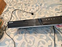 Front and top view showing disc slot, Samsung logo, attached power cable, visible heavy scratches